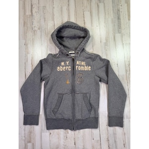 May include: A charcoal gray zip-up hoodie featuring a hood, long sleeves, and two front pockets. The chest displays the text N.Y. ATHL abercrombie in light-colored lettering, along with the number 18 in a stitched graphic design.