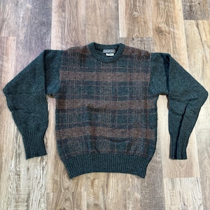 Könnte beinhalten: Ein langärmeliger Pullover mit Rundhalsausschnitt, der an den Ärmeln einfarbig dunkelgrün ist und auf der Vorderseite ein dunkelgrün-braunes Karomuster aufweist. Das Kleidungsstück hat einen gerippten Kragen, Bündchen und Saum sowie ein Etikett.