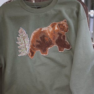 Puede incluir: Una sudadera de cuello redondo color verde oliva con un parche bordado grande y detallado en la parte delantera. El parche muestra un oso pardo caminando junto a un árbol de hoja perenne alto y texturizado. El bordado usa tonos marrones.