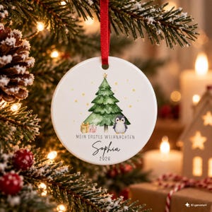 Könnte beinhalten: Ein runder, weißer Keramik-Weihnachtsbaumschmuck an einem roten Band mit einem Aquarell-Motiv aus einem grünen Tannenbaum, einem kleinen Pinguin und Geschenken. Der Text lautet "MEIN ERSTES WEIHNACHTEN", "Sophia" und "2026".