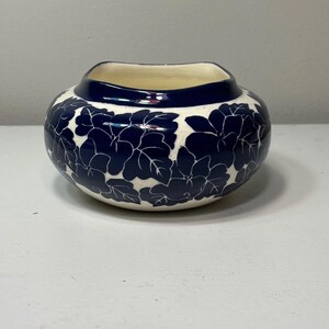 Puede incluir: Un cuenco de cerámica redondo con una base de color crema y un patrón floral azul oscuro que cubre el exterior. El cuenco presenta un borde ancho y liso de color azul oscuro y un cuerpo redondeado que se estrecha hacia la abertura, con acabado brillante.