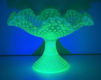 May include: A glowing neon green pedestal bowl featuring a hobnail texture and a ruffled, scalloped rim. The glass vessel emits a bright light, showcasing a raised dot pattern across the bowl and the base, with vertical ribbing on the stem.