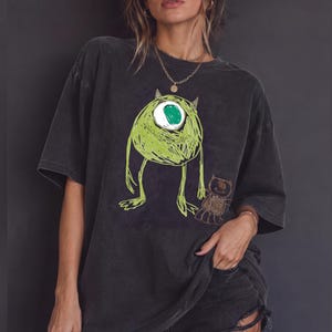 Peut inclure: Un t-shirt gris anthracite oversize orné d'une illustration verte au style griffonné représentant un monstre à un œil avec un grand œil blanc et une petite figurine brune ressemblant à un ours, au rendu brut et dessiné à la main.
