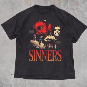 Puede incluir: Una camiseta negra de manga corta con un estampado gráfico que muestra la palabra SINNERS en letras grandes rojas y naranjas. El diseño incluye varias figuras, una pequeña cruz blanca y el texto YOU KEEP DANCING WITH THE DEVIL y ONE DAY HE'S GONNA FOLLOW YOU HOME.