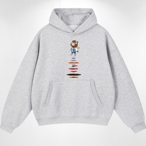 May include: A light gray hooded sweatshirt featuring a graphic of a brown bear character wearing a blue jacket and jeans, positioned above a vertical stack of colorful, circular, disc-like shapes in various hues including blue, orange, red, and black.
