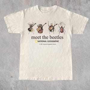 Op de afbeelding: Een crèmekleurig T-shirt met korte mouwen en een afbeelding van vier kevers die muziekinstrumenten bespelen. De tekst luidt meet the beetles, NATIONAL GEOGRAPHIC en 2001 National Geographic Society. Het ontwerp staat centraal op de borst.