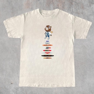 May include: A cream-colored short-sleeve t-shirt featuring a graphic of a brown bear wearing a blue jacket and jeans, positioned above a vertical stack of colorful circular vinyl records in various shades of red, blue, orange, and black.