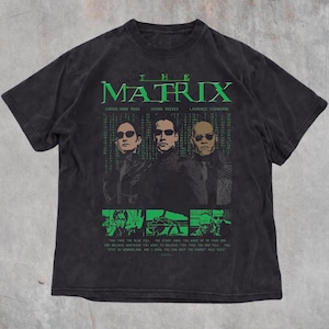 Può includere: Una t-shirt a maniche corte color nero antracite con una stampa grafica verde della locandina del film The Matrix. Il design include i ritratti di Carrie-Anne Moss, Keanu Reeves e Laurence Fishburne, con il titolo del film e il testo citato.
