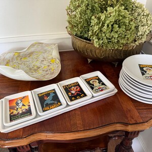 May include: A rectangular white ceramic serving tray holding three small rectangular dishes, each featuring vintage-style advertisements for Melchior, Mandarinetta Isolabella, Florio S.O.M, and Martini Vermouth, alongside a stack of white plates.