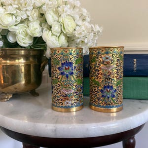 May include: Two cylindrical metal vessels featuring intricate cloisonné enamel work. The surface displays a repeating pattern of gold filigree scrolls interspersed with vibrant blue, green, pink, and white floral motifs against a metallic gold background.