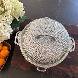 May include: A round, silver-colored metal pot with a lid, featuring a distinct hammered texture across the entire surface. The pot includes two side handles and a central handle on the lid, all crafted from the same metallic material.