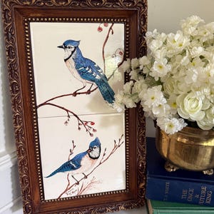 May include: A rectangular wall art piece featuring two stacked ceramic tiles with blue jay illustrations perched on branches with red berries, enclosed in an ornate, dark brown wooden frame with a beaded inner edge and carved details.