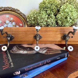 May include: A rectangular wooden wall rack featuring three brass-toned metal hooks, each accented with white ceramic knobs at the top and bottom ends. The hooks are mounted onto a polished, medium-toned wooden board with beveled edges.