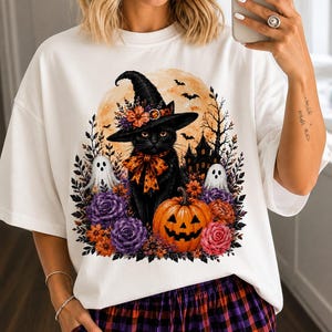 May include: A white t-shirt featuring a graphic print of a black cat wearing a witch hat and an orange bow. The design includes a jack-o'-lantern, two small ghosts, purple and orange flowers, a full moon, and a silhouette of a haunted house.
