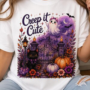 May include: A white t-shirt featuring a graphic print with the text "Creep it Cute" above a scene of a black cat with a purple bow, a ghost with a pink bow, pumpkins, flowers, a wrought iron gate, a lantern, and a purple moon with bats.