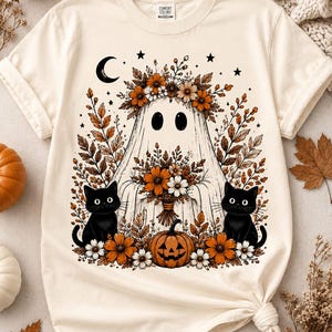 Könnte beinhalten: Ein cremefarbenes T-Shirt mit einem Grafikdruck eines Geistes, der einen Blumenkranz trägt, einen herbstlichen Blumenstrauß hält, von zwei schwarzen Katzen flankiert wird und mit einem Kürbis, einer Mondsichel und Sternen verziert ist.