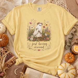 May include: A pale yellow t-shirt featuring a graphic of a ghost wearing a flower crown and holding a garden hoe, surrounded by colorful wildflowers. The text "just hoeing around" is printed below the ghost in a cursive script font.