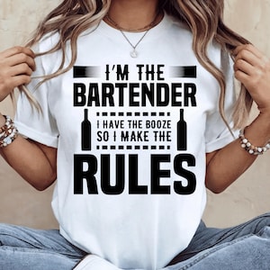 May include: A white short-sleeved t-shirt featuring black text that reads I'M THE BARTENDER I HAVE THE BOOZE SO I MAKE THE RULES. The design includes two black bottle silhouettes flanking the central text, all printed on the front of the shirt.