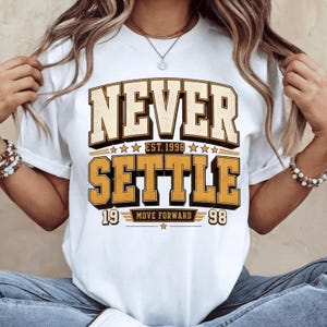May include: A white short-sleeve t-shirt featuring a graphic design with the text NEVER SETTLE in large block letters. Below, the text reads EST. 1998, MOVE FORWARD, and 19 98, accented with stars and horizontal lines in shades of yellow and black.