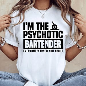 Könnte beinhalten: Ein weißes kurzärmeliges T-Shirt mit schwarzem Text, der I'M THE PSYCHOTIC BARTENDER EVERYONE WARNED YOU ABOUT lautet, begleitet von einer kleinen schwarzen Grafik eines Cocktail-Shakers über dem Wort PSYCHOTIC.