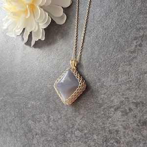 Op de afbeelding: Een ketting met een ruitvormige, gladde grijze stenen hanger, gevat in een fijn goudkleurig draadgaas, hangend aan een bijpassende dunne goudkleurige ketting. De hanger heeft een gepolijst, reflecterend oppervlak en een geometrische vorm.