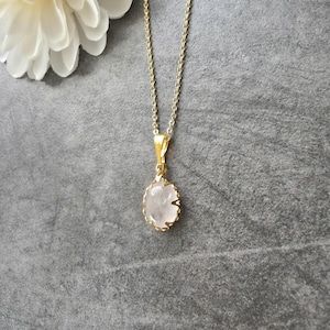 Puede incluir: Un collar de tono dorado con una cadena fina y un colgante ovalado que presenta una piedra lisa, translúcida y de color rosa pálido, engastada en un marco de metal dorado festoneado. El colgante cuelga verticalmente de una argolla sencilla.