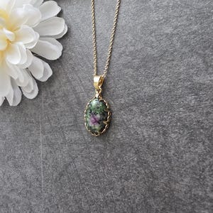 Può includere: Una collana con catenina color oro dotata di un ciondolo ovale con una pietra screziata verde e magenta. La pietra è incastonata in una montatura in metallo color oro smerlato, sospesa a una sottile catena a maglie che pende verticalmente.
