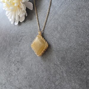 Könnte beinhalten: Ein rautenförmiger, gelber Edelsteinanhänger, der in einem feinen, goldfarbenen Drahtgeflecht eingefasst ist und an einer passenden, dünnen, goldfarbenen Metallkette hängt. Der Stein hat eine glatte, polierte Oberfläche mit feiner Marmorierung.