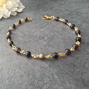 Peut inclure: Un bracelet délicat composé d'une chaîne dorée alternée de petites perles rondes marron foncé et noires. Le bijou présente une perle centrale dans un ton gris plus clair et est fermé par un fermoir mousqueton de couleur dorée.
