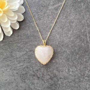 Op de afbeelding: Een hartvormige, lichtroze stenen hanger omhuld door een delicaat goudkleurig draad, hangend aan een dunne goudkleurige metalen ketting. De hanger heeft een glad, gepolijst oppervlak en is aan de ketting bevestigd met een kleine goudkleurige ring.