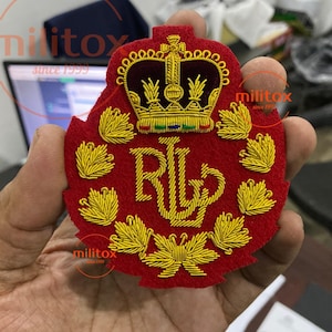 以下が含まれることがあります： 金色の刺繍糸で装飾された赤いフェルト製のワッペン。中央には王冠とRLの文字が配置され、その周囲を金色の刺繍で施された葉が円状に囲んでいます。ワッペンは縁が波型にカットされており、金属のような質感の仕上がりになっています。