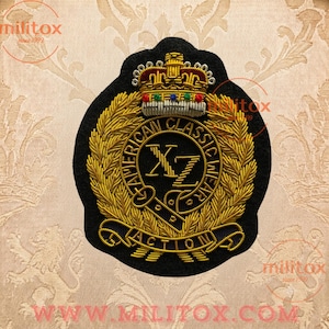 May include: A black embroidered patch featuring a gold crown with red, blue, and green accents at the top. The centre displays the letters XZ surrounded by a circular border reading AMERICAN CLASSIC WEAR, framed by gold laurel leaves and a banner reading ACTION.