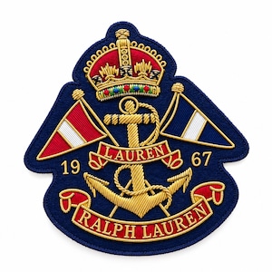 Puede incluir: Un parche bordado azul marino con un ancla dorada, una corona y dos banderas. El texto LAUREN aparece en una cinta roja sobre el ancla, y RALPH LAUREN en una cinta roja en la parte inferior. Los números 19 y 67 están a ambos lados.