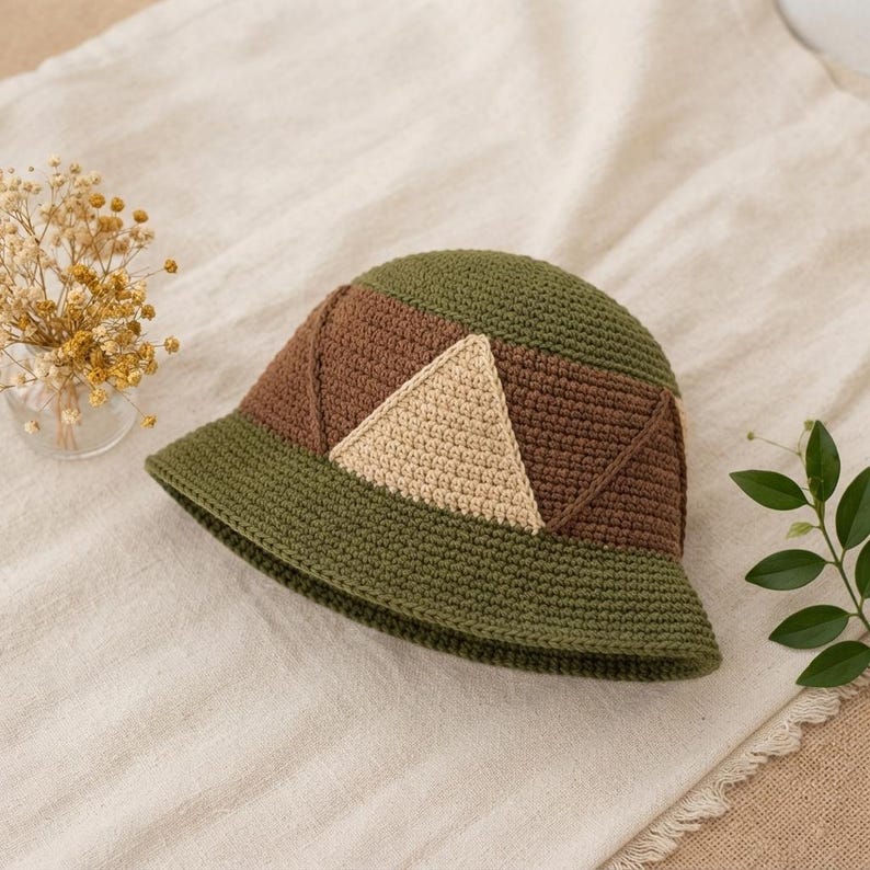 May include: A crochet bucket hat featuring a color-blocked design with olive green, dark brown, and cream sections. The hat is constructed with a dense, textured stitch pattern, showcasing a prominent cream-colored triangle on the side panel.