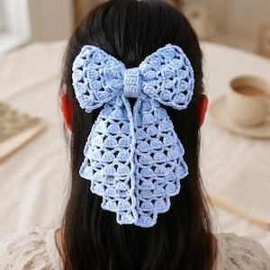 May include: A light blue crochet hair bow featuring an intricate openwork pattern with a central knot and two long, cascading tails. The accessory is constructed from yarn with a repeating geometric stitch design, creating a textured appearance.