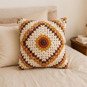 Könnte beinhalten: Ein quadratisches Häkelkissen mit einem kreisförmigen Granny-Square-Muster in der Mitte. Das Design zeigt konzentrische Ringe aus dunkelbraunem, rostorangem, senfgelbem und cremefarbenem Garn, die ein strukturiertes, geometrisches Blumenmotiv bilden.
