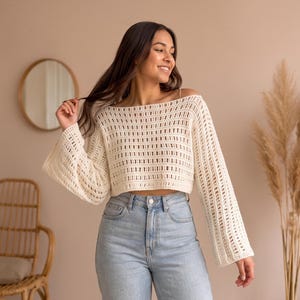 May include: A cream-coloured, long-sleeved, cropped crochet jumper featuring an open-knit vertical stripe pattern, a wide boat neckline, and loose, flared sleeves. The garment is paired with light blue denim jeans with a visible metal button.