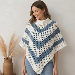 Peut inclure: Un poncho en crochet doté d'un col montant et d'un motif en V alternant des bandes de laine blanche et bleu cendré. Le vêtement est réalisé avec un point de granny square classique, créant un aspect texturé et ajouré qui tombe sur les épaules et le torse avec un ourlet en pointe.