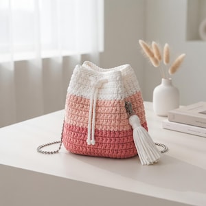 May include: A crochet bucket bag featuring a color-blocked design in white, light pink, and dark pink. The bag includes a white drawstring closure, a silver-toned metal chain strap, and a large white tassel attached to the side with a metal clip.