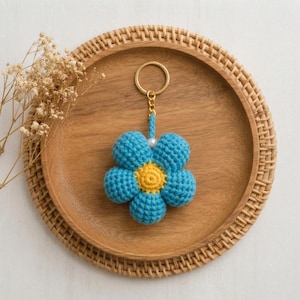 Peut inclure: Un porte-clés fleur en crochet composé de cinq pétales bleus arrondis et d'un centre jaune texturé. La fleur est fixée à un anneau métallique doré par un cordon bleu court orné d'une petite perle blanche, formant un accessoire doux au toucher.