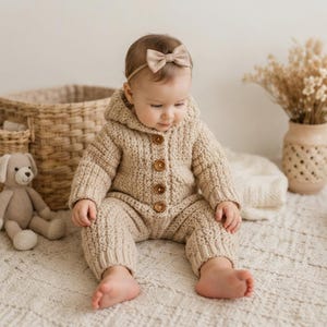 May include: A beige crocheted hooded romper with long sleeves and four brown wooden buttons down the front. The garment features a textured knit pattern and ribbed cuffs at the wrists and ankles. A matching beige bow headband is worn on the head.