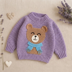 May include: A lavender long-sleeve crochet sweater featuring a central brown bear applique with a light blue scarf. The sweater has a ribbed crew neckline, cuffs, and hem, showcasing a textured yarn construction with a consistent stitch pattern.
