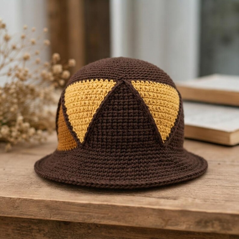 May include: A crochet bucket hat featuring a dark brown base color with contrasting mustard yellow triangular patterns integrated into the crown. The hat has a rounded top and a wide, flat brim, all constructed with a consistent yarn stitch.