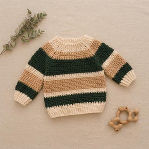 Könnte beinhalten: Ein langärmeliger Häkelpullover mit horizontalen Streifen in Creme, Beige und dunklem Waldgrün. Das Kleidungsstück hat einen Rundhalsausschnitt und gerippte Bündchen und zeigt eine strukturierte Garnkonstruktion mit einem gleichmäßigen Muster.