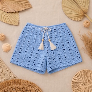 Può includere: Pantaloncini all'uncinetto azzurri con motivo traforato verticale, orlo smerlato e vita con coulisse dotata di due cordini bianchi che terminano con perline di legno e nappe color crema. Il capo è realizzato in filato di colore blu tinta unita.