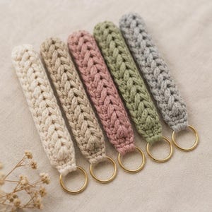 Könnte beinhalten: Fünf gehäkelte Schlüsselanhänger in einer Reihe, jeder mit einer dicken geflochtenen Textur in Creme, Beige, Altrosa, Salbeigrün und Hellgrau. Jeder Schlüsselanhänger ist am unteren Ende mit einem runden goldfarbenen Metallring versehen.
