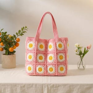 May include: A pink crochet tote bag constructed from square panels, each featuring a central yellow daisy with white petals. The bag includes two matching pink shoulder straps and a textured, handmade appearance with a consistent grid pattern.