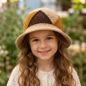 May include: A crochet bucket hat featuring a geometric color-blocked design in shades of beige, mustard yellow, and dark brown. The hat has a rounded crown and a downward-sloping brim, constructed with a dense, textured stitch pattern throughout.