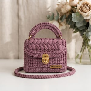 Può includere: Una borsa all'uncinetto color malva con una spessa patta intrecciata, una chiusura a scatto in metallo dorato e una tracolla abbinata. Una piccola etichetta rettangolare in legno con la scritta "Handmade" è fissata sulla parte anteriore.