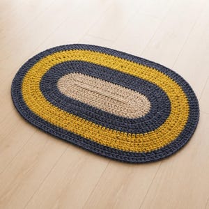Puede incluir: Una alfombra de ganchillo de forma ovalada con bandas concéntricas de color. El centro es un óvalo beige sólido, rodeado por un anillo grueso gris oscuro, seguido de un borde amarillo mostaza vibrante y un borde exterior de hilo gris oscuro.